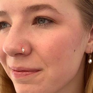 May include: A close-up of a person's face with a small pearl nose ring and a pearl earring. The person has blonde hair and is wearing makeup.