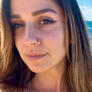 May include: A close-up of a person's face, with a silver nose ring featuring a small, round, red gemstone. The person has brown hair and a gold necklace. The background is a blurred view of the sea and sky.