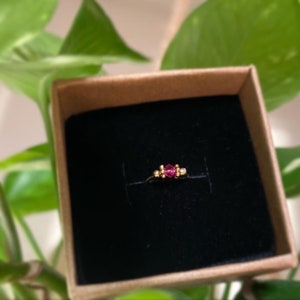 May include: A gold-colored ring with a central, faceted, pink gemstone and smaller, round, gold-colored accents on either side. The ring is presented in a small, open, brown box with a black interior, set against a backdrop of green foliage.