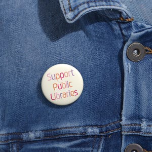 Può includere: Una spilla rotonda con sfondo color crema che riporta la scritta Support Public Libraries in lettere multicolori. La spilla è fissata su una giacca di jeans blu con cuciture visibili e un bottone in metallo scuro.
