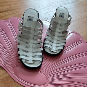 May include: A pair of white leather sandals with a caged design. The sandals have a chunky heel and are sitting on a pink and white patterned surface.