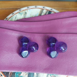 May include: A pair of vintage purple earrings with a three-orb design. The earrings are set on a deep purple leather surface. The background includes a decorative plate and a wooden surface.