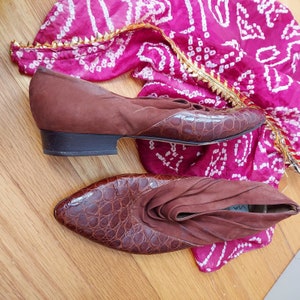 A pair of brown leather shoes with a crocodile pattern at the vamp and toe. The shoes have an almond pointed toe and a low stacked heel. The ruched cuff adds a look of distinction, tailored by Vig Spiga of Italy. The shoes are displayed on a pink and white patterned fabric.