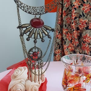 May include: An ornate, antique-style necklace with a large oval red gemstone and intricate metalwork. The necklace features multiple chains and a circular pendant with a smaller red stone. The design is reminiscent of vintage jewelry.