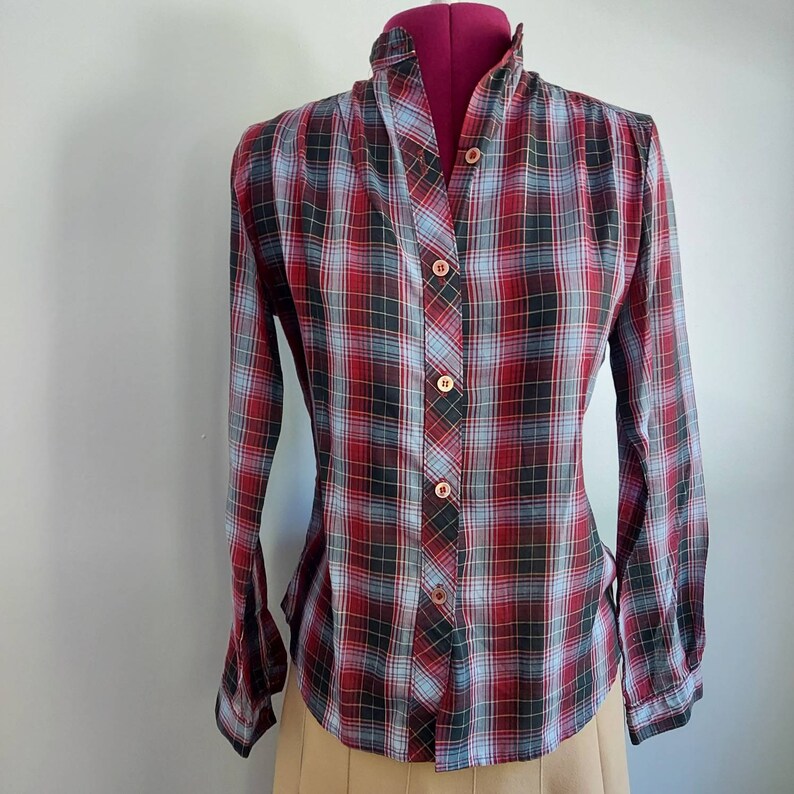 May include: A long-sleeved plaid shirt in red, black, and light blue, buttoned up. The shirt is displayed on a mannequin. The plaid pattern is a classic design, and the shirt has a high collar.