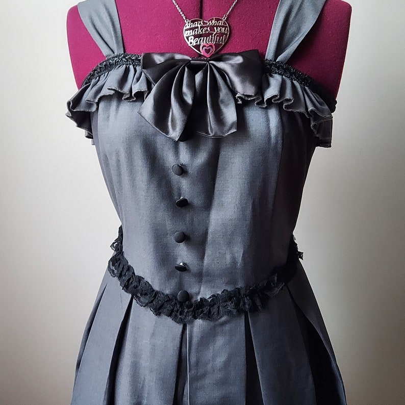 Op de afbeelding: Een grijze, mouwloze jurk met een zwarte kanten rand en een zwarte strik bij de halslijn. De jurk heeft een zilveren ketting met een hartvormige hanger waarop "That's what makes you beautiful" staat.