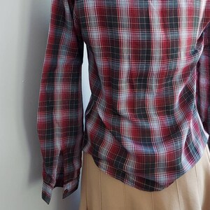 May include: A long-sleeved plaid shirt in a classic pattern of red, black, and gray. The shirt is buttoned and has a tailored fit. The bottom of the shirt is visible, paired with a tan skirt.