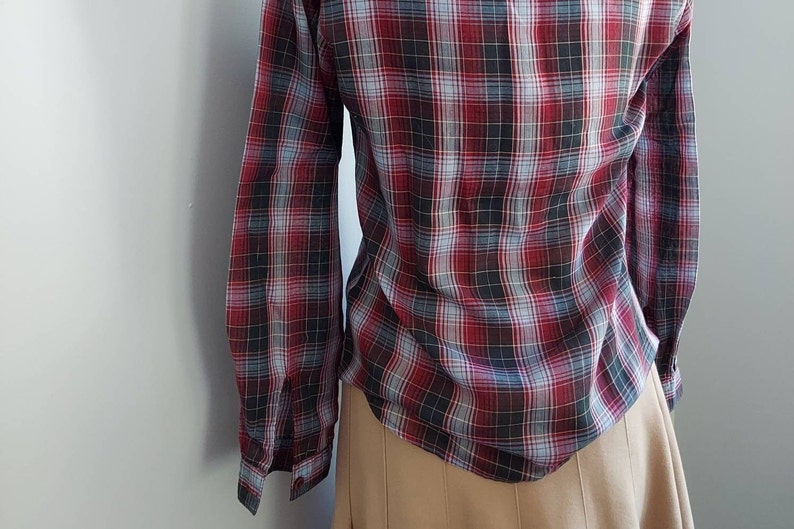 May include: A long-sleeved plaid shirt in a classic pattern of red, black, and gray. The shirt is buttoned and has a tailored fit. The bottom of the shirt is visible, paired with a tan skirt.