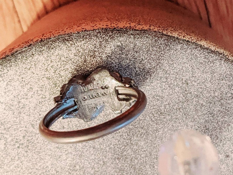 May include: A silver-toned metal ring featuring a thin, circular band and a textured, raised centerpiece. The underside of the centerpiece displays the stamped text "AVON" with a copyright symbol, indicating the manufacturer of this jewelry piece.