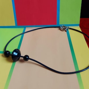 May include: A black cord necklace featuring three black beads, with a larger disc-shaped bead centered between two smaller spherical beads. The necklace is secured with a silver-toned metal toggle clasp at the back, providing a simple and functional design.