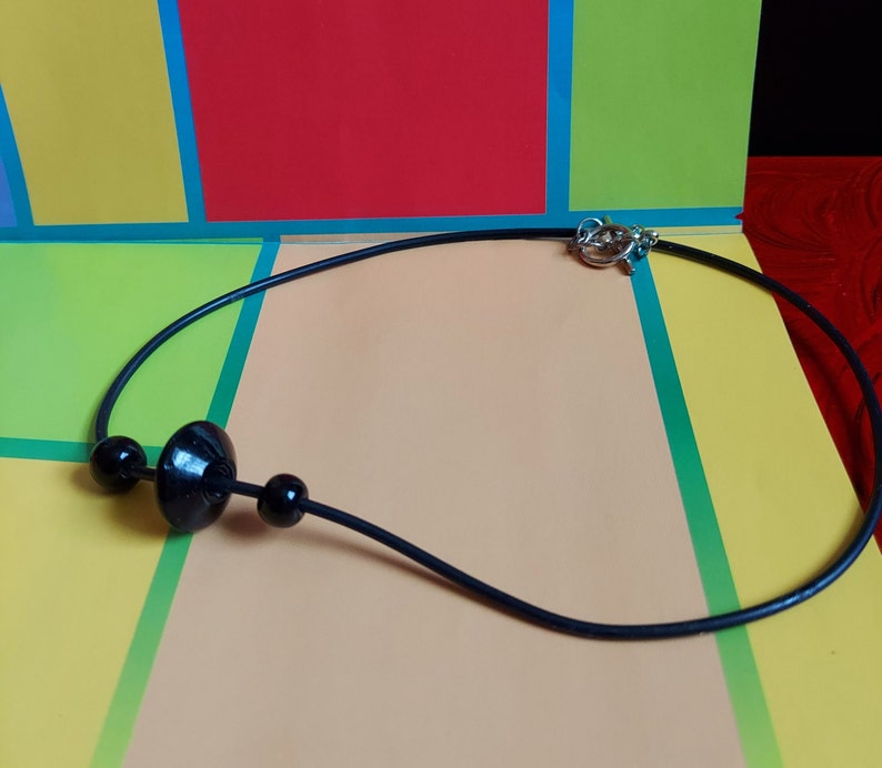 May include: A black cord necklace featuring three black beads, with a larger disc-shaped bead centered between two smaller spherical beads. The necklace is secured with a silver-toned metal toggle clasp at the back, providing a simple and functional design.