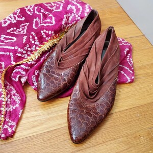 A pair of brown leather mules in a croc pattern, with a ruched suede vamp. The toes are pointed almond style, and the heels are low, stacked.The shoes are displayed on a wooden surface with a pink and white patterned fabric in the background.