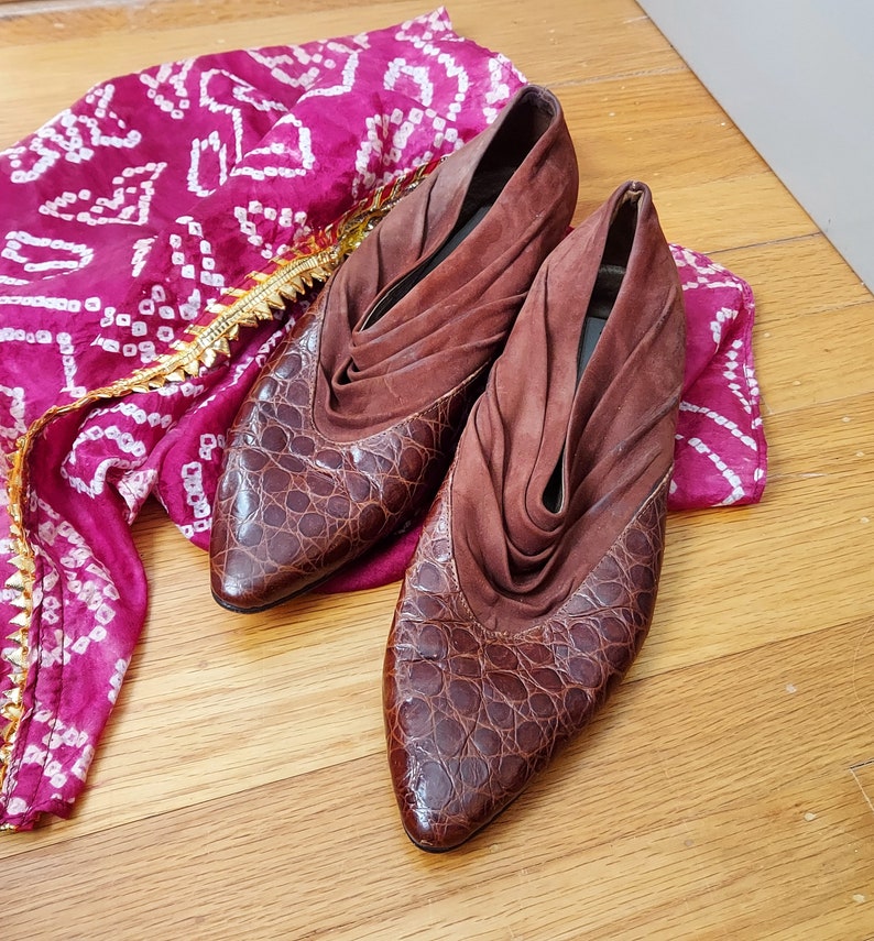 A pair of brown leather mules in a croc pattern, with a ruched suede vamp. The toes are pointed almond style, and the heels are low, stacked.The shoes are displayed on a wooden surface with a pink and white patterned fabric in the background.