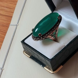May include: A green oval gemstone ring with a silver filigree setting. The ring is in a green velvet ring box.