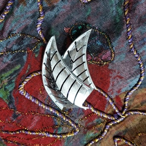 Puede incluir: Un broche de plata en forma de hoja con un diseño texturizado, sobre un fondo de tela colorido.