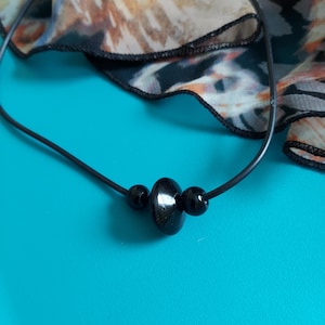 May include: A black cord necklace featuring a central, flattened, circular black bead flanked by two smaller, spherical black beads. The necklace is displayed against a turquoise background with a patterned, semi-sheer fabric visible at the top.