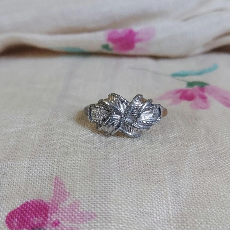 May include: A silver-toned metal ring featuring a central knot design. The surface of the knot displays a textured, brushed finish with fine horizontal lines and small beaded edges along the borders of the intertwined metal loops.