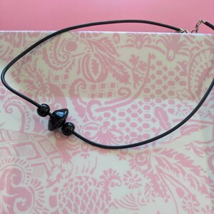 May include: A black cord necklace featuring three central black beads, including two spherical beads flanking a single wider, disc-shaped bead. The necklace includes a silver-toned metal clasp mechanism at the end of the black cord.