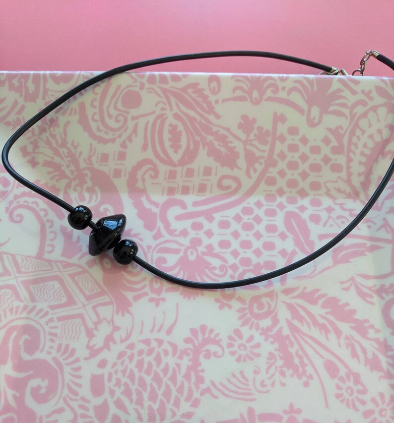 May include: A black cord necklace featuring three central black beads, including two spherical beads flanking a single wider, disc-shaped bead. The necklace includes a silver-toned metal clasp mechanism at the end of the black cord.