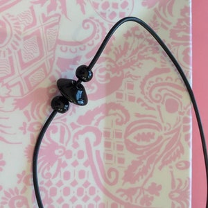 May include: A black cord necklace featuring three glossy black beads. The central bead is shaped like a flattened disc, flanked by two smaller spherical beads. The cord is thin, flexible, and dark, providing a stark contrast to the smooth, reflective surfaces of the black beads strung along its length.