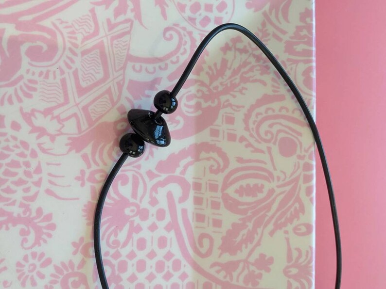 May include: A black cord necklace featuring three glossy black beads. The central bead is shaped like a flattened disc, flanked by two smaller spherical beads. The cord is thin, flexible, and dark, providing a stark contrast to the smooth, reflective surfaces of the black beads strung along its length.