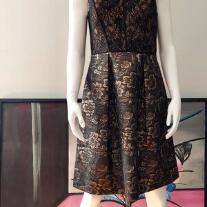 May include: Sleeveless black dress with a floral pattern in gold. The bodice is black lace, and the skirt is pleated. The dress is displayed on a mannequin. The dress is knee-length.