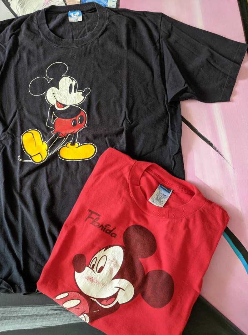 以下が含まれることがあります： ミッキーマウスがプリントされたTシャツ2枚。上のTシャツは黒地に白のミッキーマウスのプリント。下のTシャツは赤地に白のミッキーマウスのプリントで、前面に「フロリダ」の文字がプリントされている。