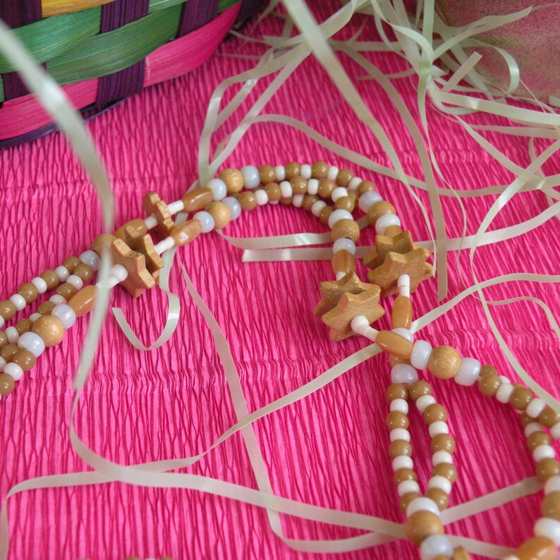 K&ouml;nnte beinhalten: Eine wei&szlig;e und braune Perlenkette mit Holzstern-Anh&auml;ngern. Die Kette liegt auf einer rosa, gekr&auml;uselten Oberfl&auml;che mit wei&szlig;em Band.