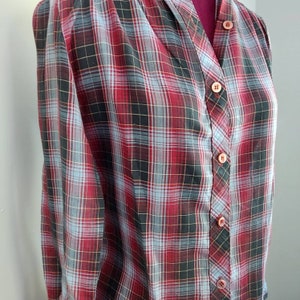 May include: A vintage plaid button-down shirt in a red, black, and light blue pattern. The shirt has a V-neck and is buttoned up the front with matching buttons. The fabric appears to be lightweight and suitable for warmer weather.