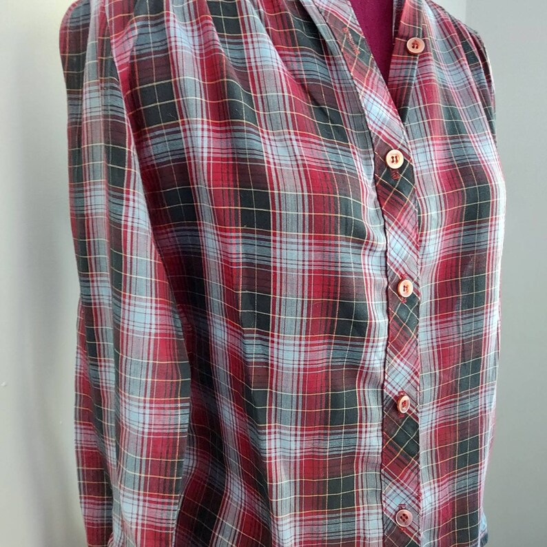 May include: A vintage plaid button-down shirt in a red, black, and light blue pattern. The shirt has a V-neck and is buttoned up the front with matching buttons. The fabric appears to be lightweight and suitable for warmer weather.