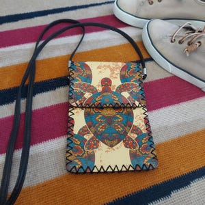Puede incluir: Un pequeño bolso bandolera rectangular con una correa negra. El bolso presenta un diseño de tortuga colorido en tonos azules, rojos y amarillos sobre un fondo crema. Los bordes están cosidos con hilo negro. Un par de zapatillas grises están en el fondo.