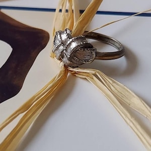 May include: A silver-toned metal ring featuring a central knot design with a textured, brushed finish. The band is smooth and circular, and the knot element displays intricate, fine vertical lines across its surface, creating a detailed appearance.