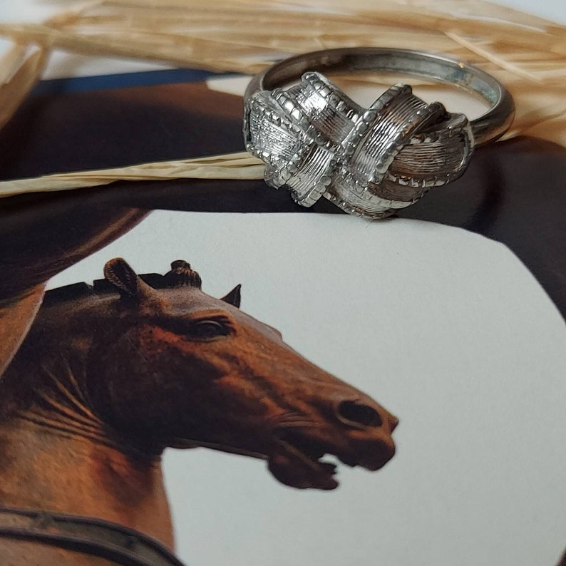 May include: A silver-toned metal ring featuring a central knot design with a textured, brushed finish. The band is smooth and circular, showcasing a detailed, woven-like pattern on the top section that creates a dimensional, layered appearance.