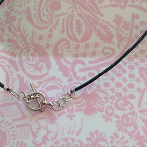 May include: A black cord necklace featuring a silver-toned toggle clasp closure. The necklace includes a circular ring and a T-bar component made of metal, connected to the black cord with small silver-toned metal crimp ends and jump rings.