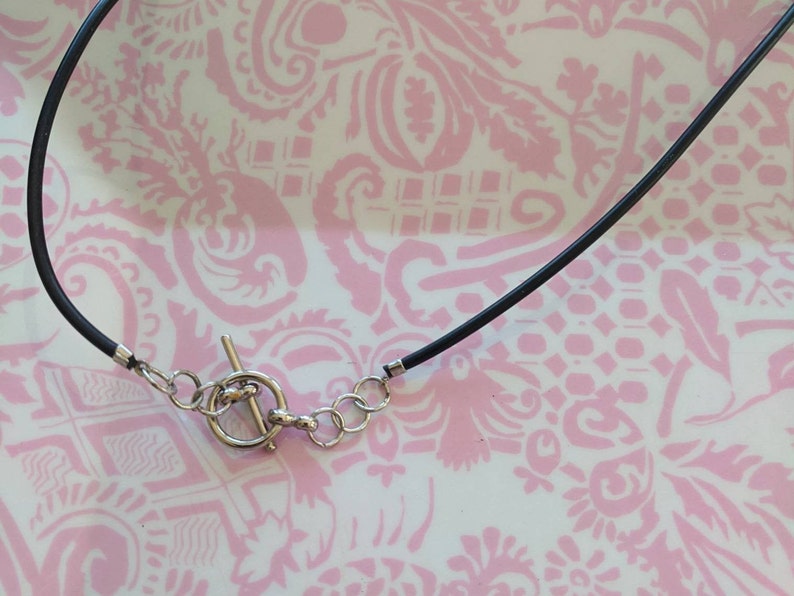 May include: A black cord necklace featuring a silver-toned toggle clasp closure. The necklace includes a circular ring and a T-bar component made of metal, connected to the black cord with small silver-toned metal crimp ends and jump rings.