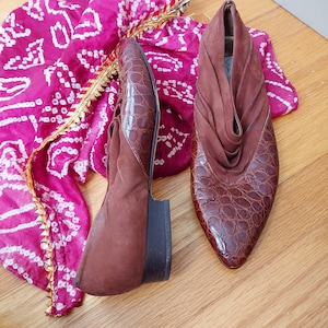 A pair of brown leather shoes with a crocodile pattern. These mule style slip-ons are depicted here, with one shoe turned to the side to show the heel height. Both mules have a vamp of ruched suede which wraps to the back of the shoe. These Vintage size 8.5 mules are displayed on a pink and white patterned fabric.