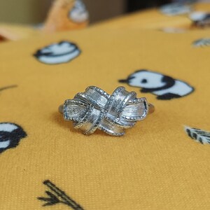 May include: A silver-toned ring featuring a central knot design with a textured, ribbed surface. The knot is composed of multiple overlapping loops that create a dimensional, woven appearance. The metal has a polished finish with fine detailing.