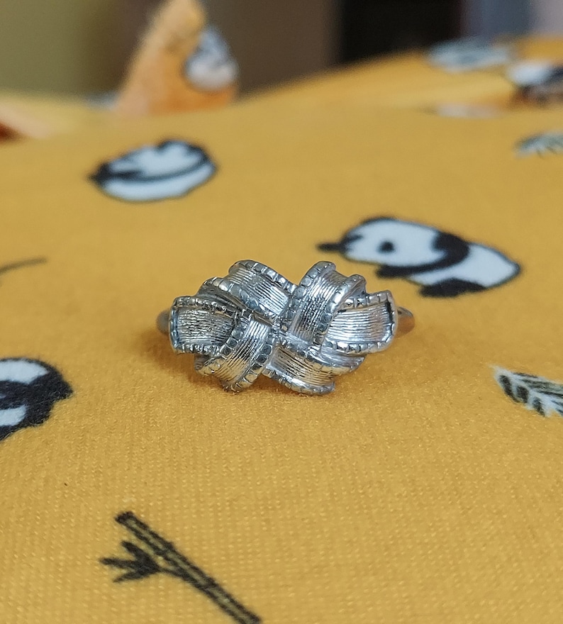 May include: A silver-toned ring featuring a central knot design with a textured, ribbed surface. The knot is composed of multiple overlapping loops that create a dimensional, woven appearance. The metal has a polished finish with fine detailing.