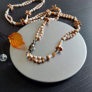 K&ouml;nnte beinhalten: Eine zweireihige Halskette aus braunen und wei&szlig;en Perlen mit einem kleinen Blumenanh&auml;nger und einem Metallverschluss.