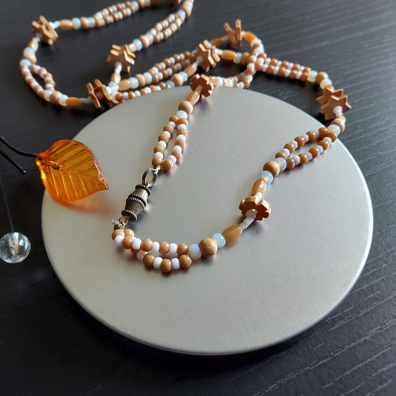 K&ouml;nnte beinhalten: Eine zweireihige Halskette aus braunen und wei&szlig;en Perlen mit einem kleinen Blumenanh&auml;nger und einem Metallverschluss.