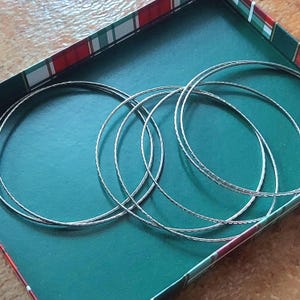 Peut inclure: Plusieurs bracelets fins de couleur argentée sont disposés dans une boîte verte avec des bordures rouges et blanches. Les bracelets ont un motif texturé et sont de forme circulaire.