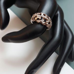 Puede incluir: Un anillo de plata con un diseño texturizado y calado. El anillo está hecho de una serie de formas superpuestas y conectadas.