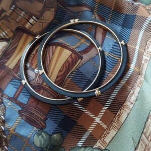 May include: Two circular bangles featuring a matte black outer band and a smooth gold-toned inner rim. Each bangle is accented with four small, evenly spaced gold-toned metal decorative elements that bridge the black and gold sections of the jewelry.