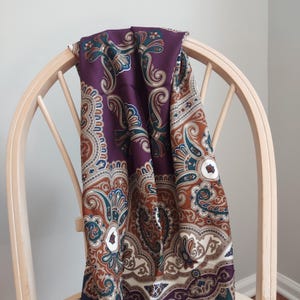 May include: A square scarf featuring a detailed paisley pattern in shades of deep purple, burnt orange, teal, and cream. The intricate design includes swirling motifs and floral-inspired shapes arranged in a symmetrical layout across the fabric.