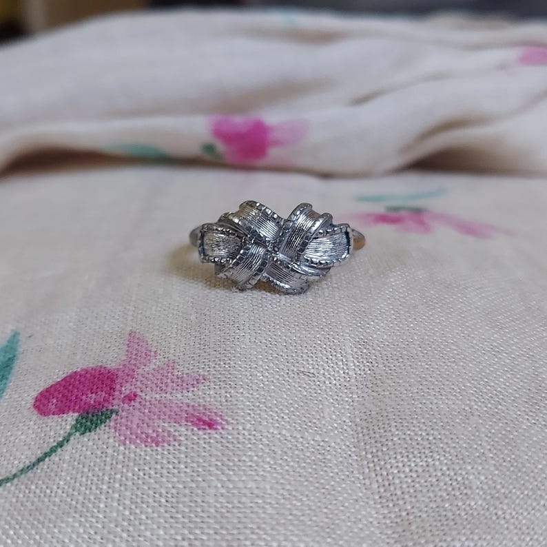 May include: A silver-toned metal ring featuring a central knot design with a textured, brushed finish. The knot is composed of interwoven bands that create a layered, dimensional appearance across the front of the ring band.