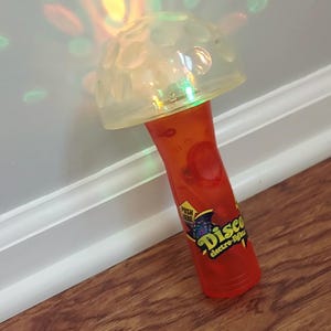 May include: A red and clear plastic disco light toy with a clear dome top. The handle has the words "Disco electro-lights" printed on it. The toy is angled on a wooden surface.