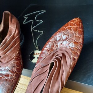A pair of two brown leather mules with a crocodile pattern. These Chelsea style shoes have a ruched suede cuff, an almond toe, and a low, stacked heel. A silver necklace with a pendant featuring a woman wearing glasses is in the background.