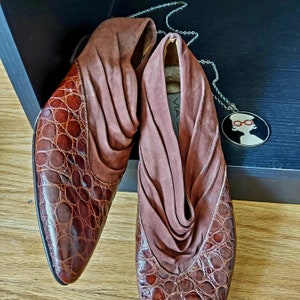 A pair of brown leather shoes with a crocodile pattern. These mule style slip-ons have a pointed almond toe and a low stacked heel. The shoes are made of a soft, supple leather, accented by a cuff of ruched suede for the vamp. Shoes are displayed on a black wooden box, with a round pendant of red, black and white nearby. These mules could be considered as Rockabilly, Western theme.