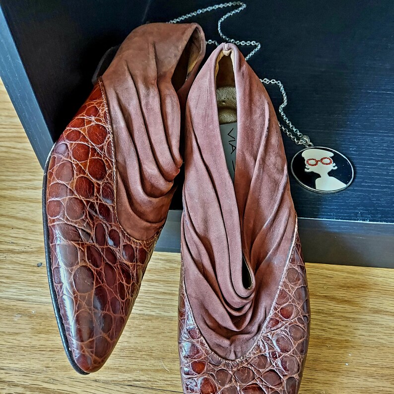 A pair of brown leather shoes with a crocodile pattern. These mule style slip-ons have a pointed almond toe and a low stacked heel. The shoes are made of a soft, supple leather, accented by a cuff of ruched suede for the vamp. Shoes are displayed on a black wooden box, with a round pendant of red, black and white nearby. These mules could be considered as Rockabilly, Western theme.