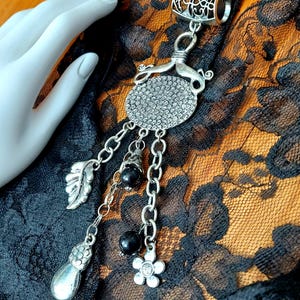 May include: Silver hair clip with a round, ornate design and dangling chains with black beads and silver charms, including a leaf, a flower, and a pear-shaped charm.